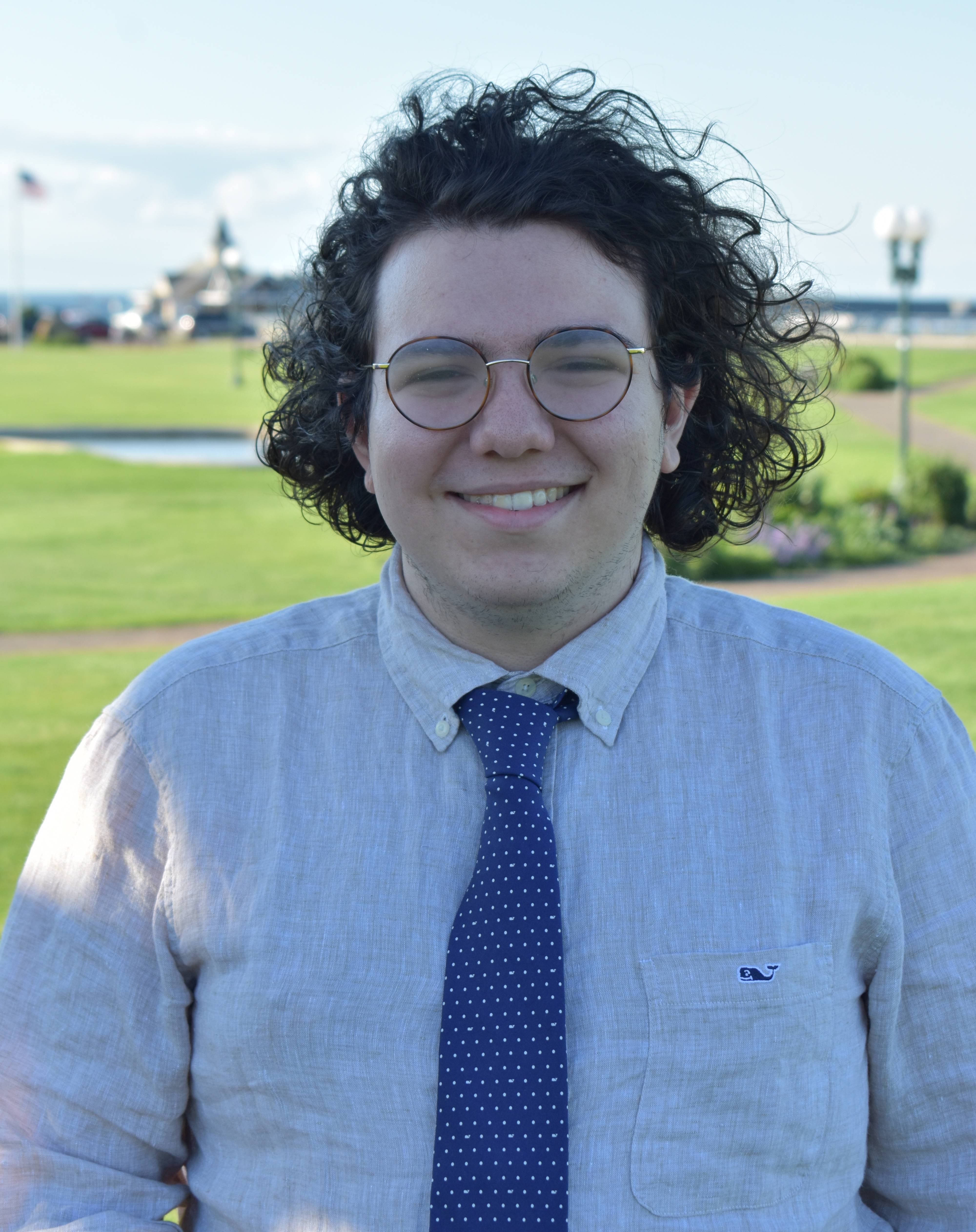 A picture of Alex Kaplan, a smiling man with glasses and curly black hair. He is wearing an off-white linen shirt and a blue tie with white dots. He stands in front of a lush park in Oak Bluffs, Massachusetts, with the ocean in the distance.