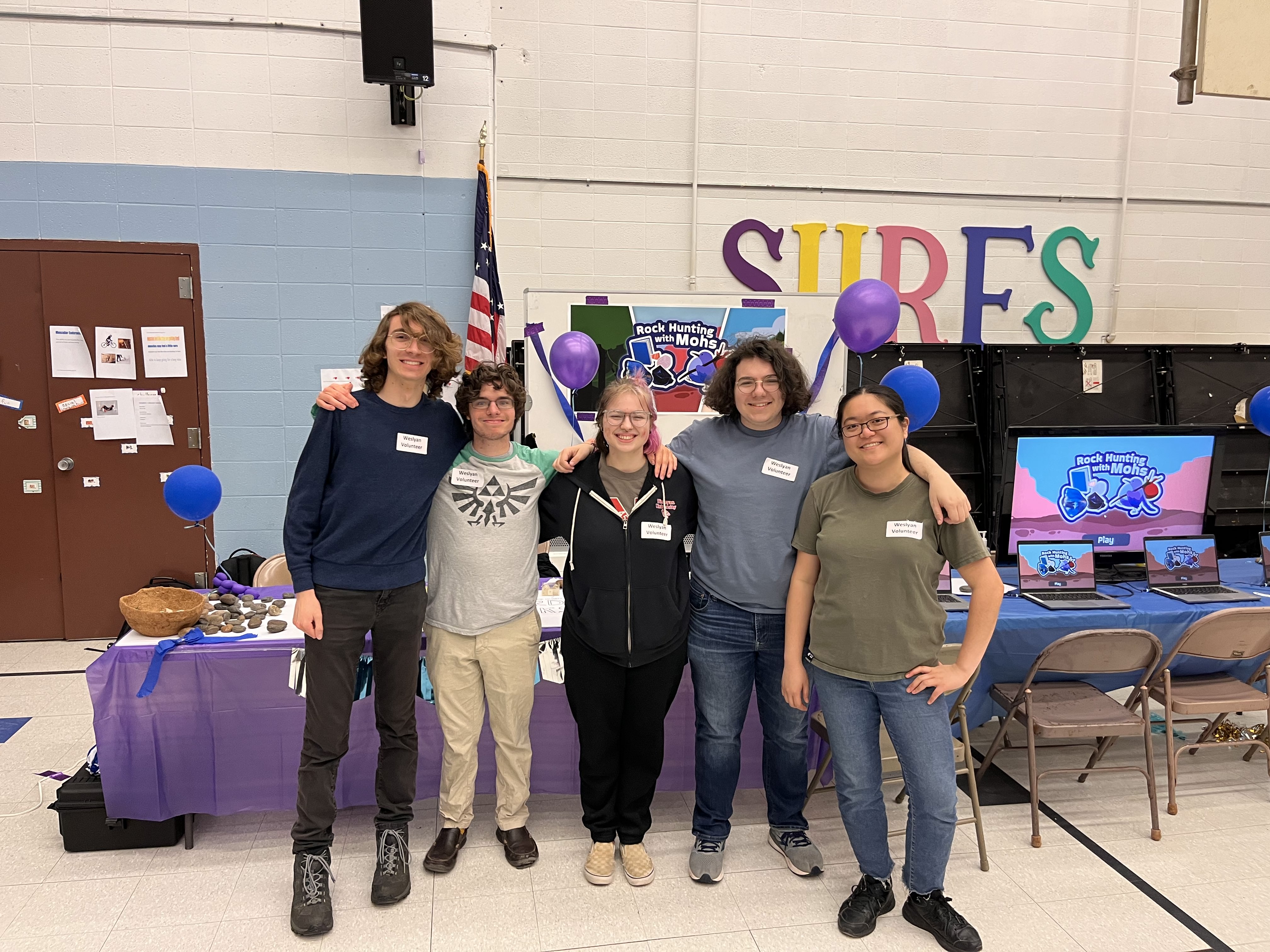 A photo of the Rock Hunting with Mohs team, standing in front of the game's carnival booth at MacDonough elementary school. They have their arms around each other's shoulders, and are smiling.